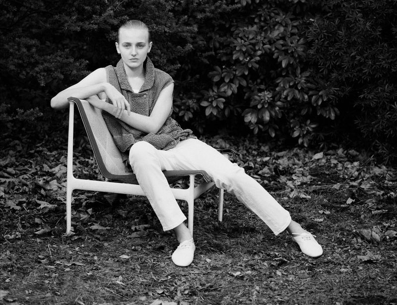 Model seated in modern chair outdoors wearing sleeveless textured top and white trousers against foliage
