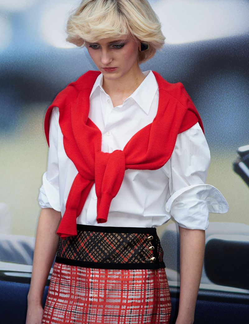 Model with blonde hair wearing a white shirt, red sweater tied around shoulders, and plaid skirt
