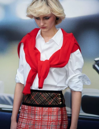 Model with blonde hair wearing a white shirt, red sweater tied around shoulders, and plaid skirt