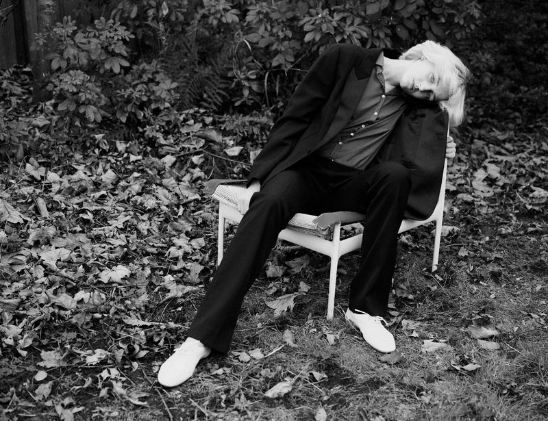 Model George Anderson seated on a white chair outdoors in a garden wearing a black blazer and trousers for Pop Magazine photographed by Charlotte Stouvenot.