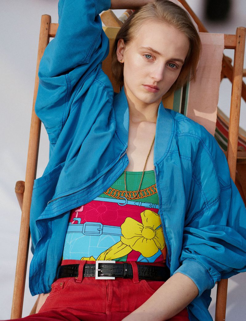 Model wearing a blue jacket and red trousers seated in a wooden deckchair for a Pop Magazine editorial shoot.