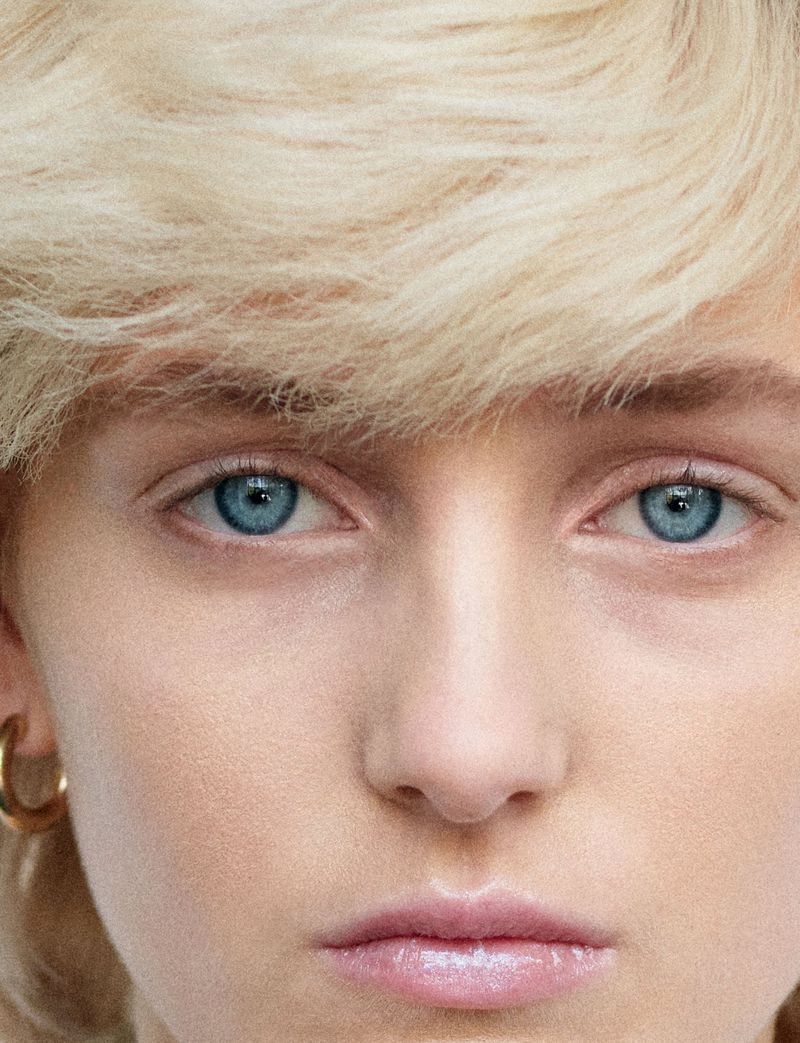 Close-up portrait of a model with short blonde hair and striking blue eyes wearing a small hoop earring.