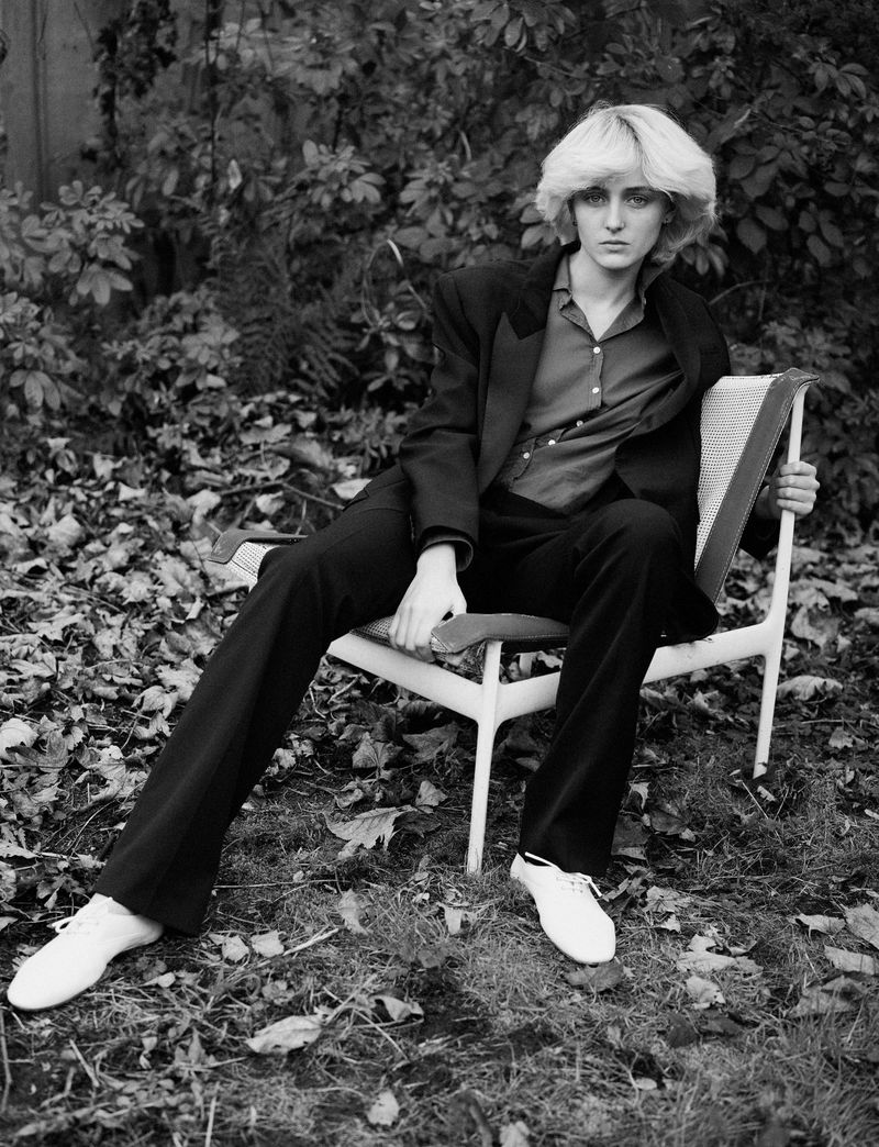 Black and white portrait of an adult woman with short blonde hair, wearing a dark jacket and pants, seated in a chair outdoors.