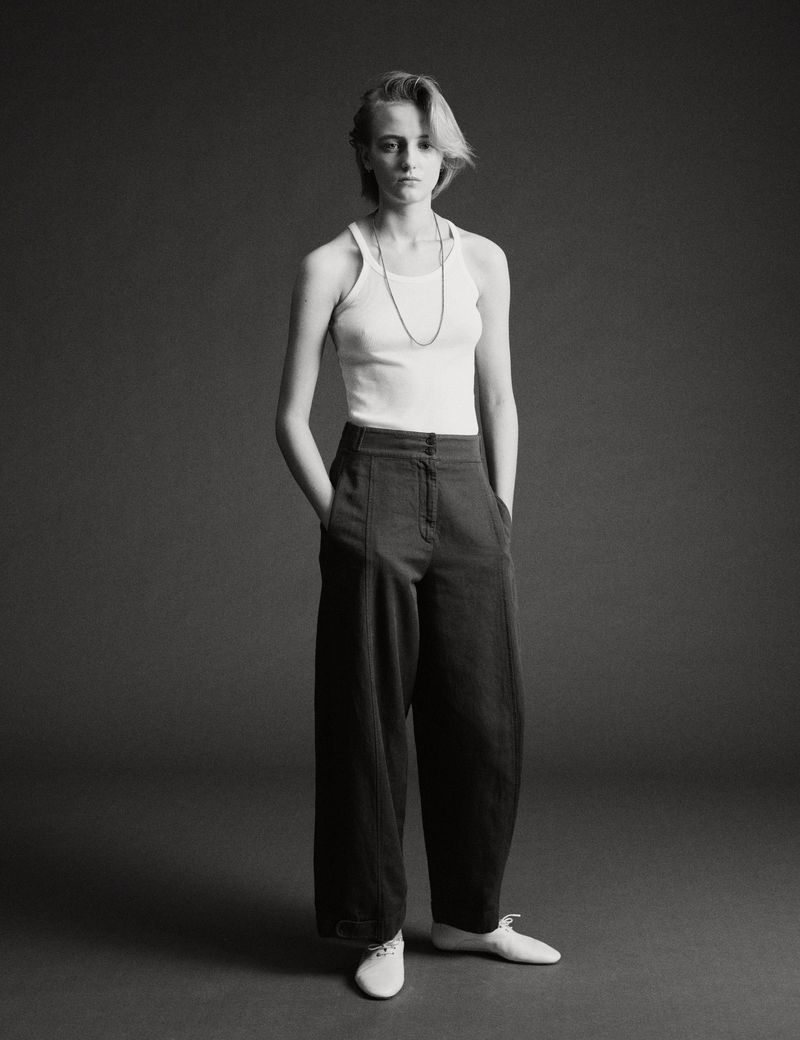 Model standing against a plain dark grey studio backdrop wearing a white tank top and dark trousers with hands in pockets.
