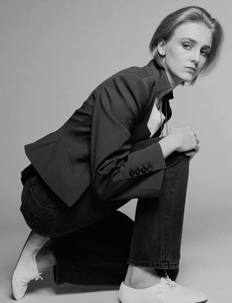 Model George Anderson crouches in a dark blazer, jeans, and white shoes against a plain grey seamless backdrop.
