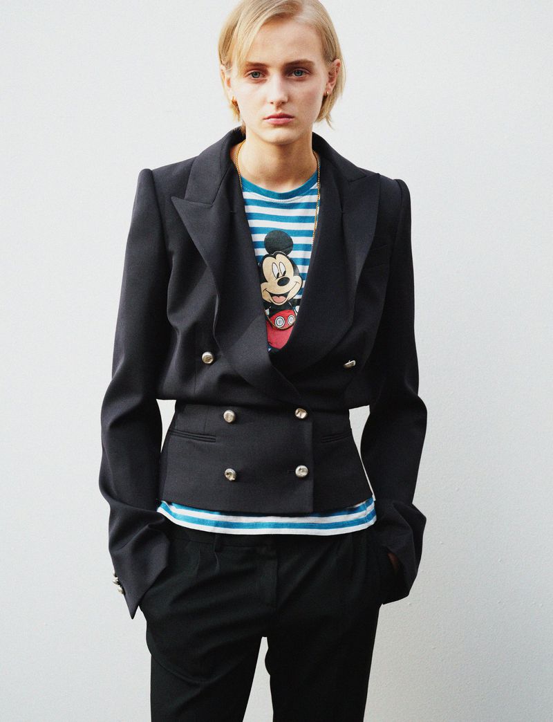 Young woman in a black blazer and striped t-shirt poses against a white wall. Studio shot of a person in business casual fashion.