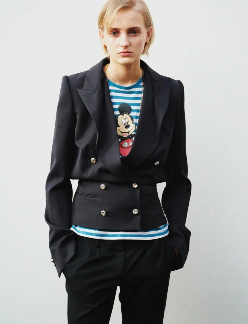 Young woman in a black blazer and striped t-shirt poses against a white wall. Studio shot of a person in business casual fashion.
