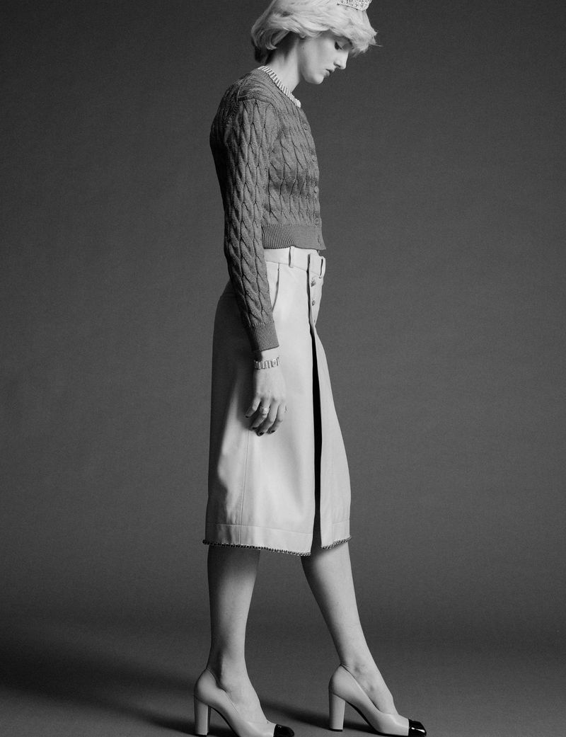 Monochrome full-length portrait of a model in a cable-knit cardigan and skirt against a dark background.