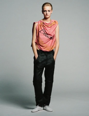 Model standing against a neutral grey backdrop wearing a pink draped top and dark trousers.