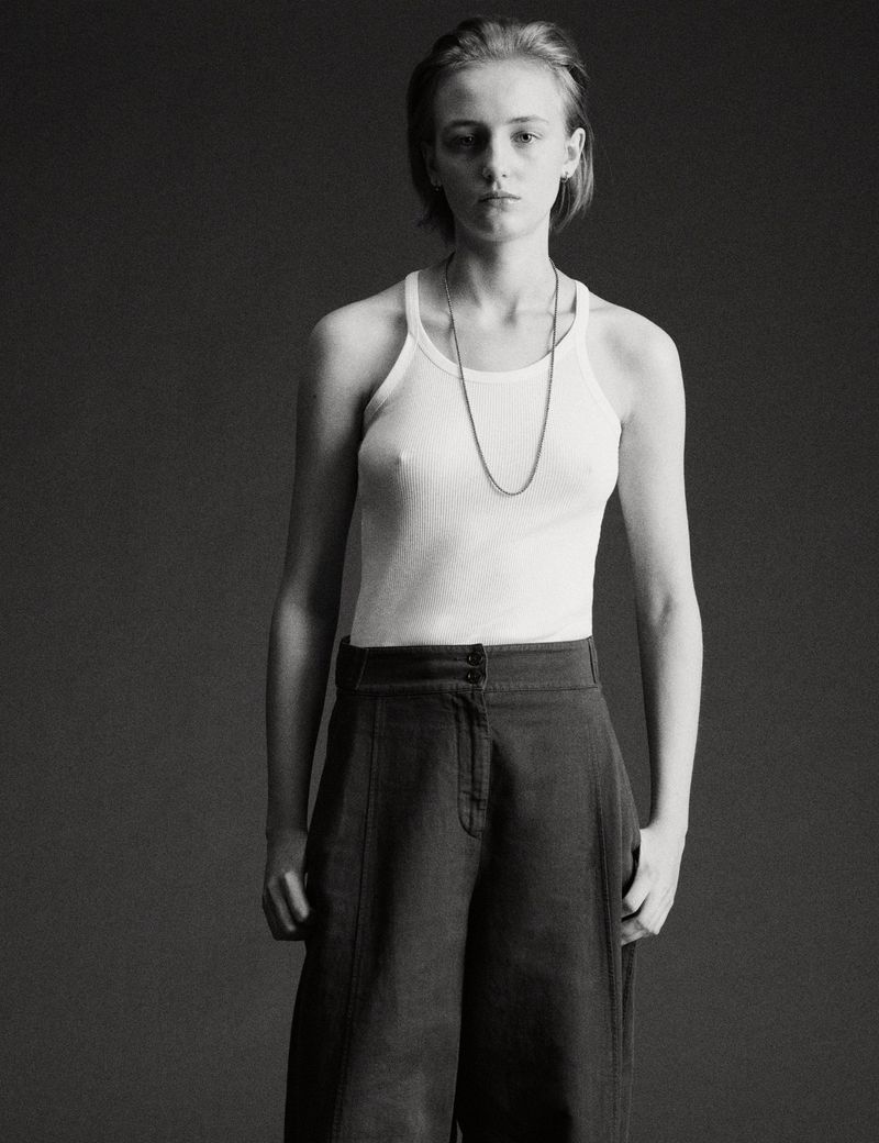 Portrait of a model wearing a white tank top and dark trousers against a plain dark studio background.