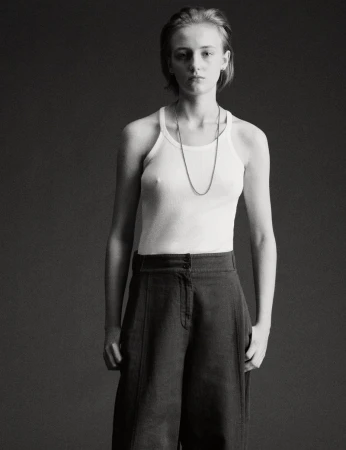 Portrait of a model wearing a white tank top and dark trousers against a plain dark studio background.