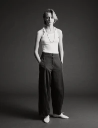 Model standing against a plain dark grey studio backdrop wearing a white tank top and dark trousers with hands in pockets.