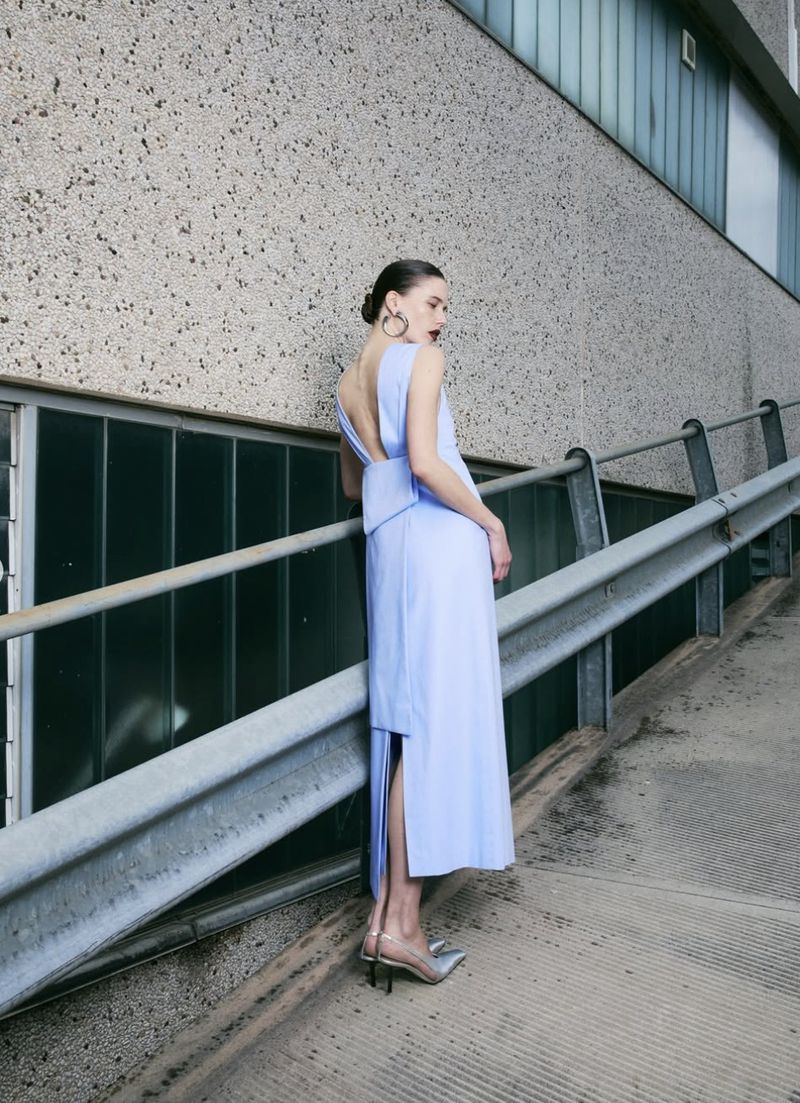 Woman in a periwinkle dress stands on a concrete incline, accessorized with hoop earrings and clear heels, adding to the minimalist aesthetic.