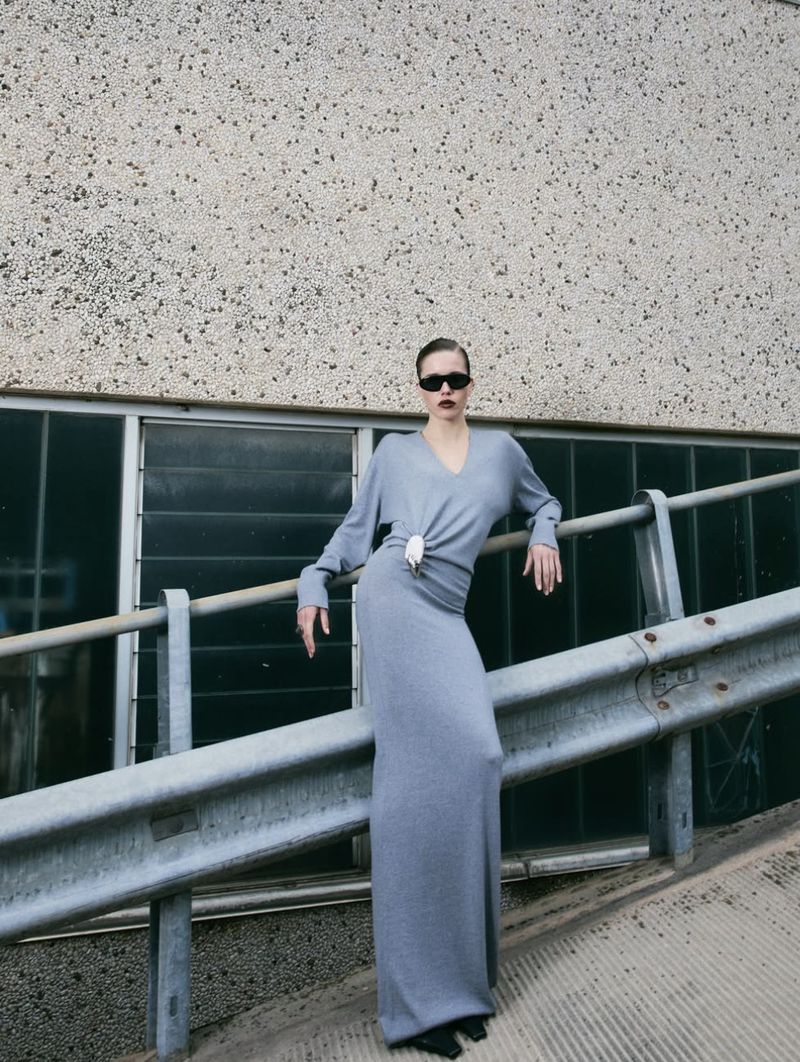 A woman wearing sunglasses and a gray dress stands on an angled ramp near a railing and a wall with dark horizontal rectangles.