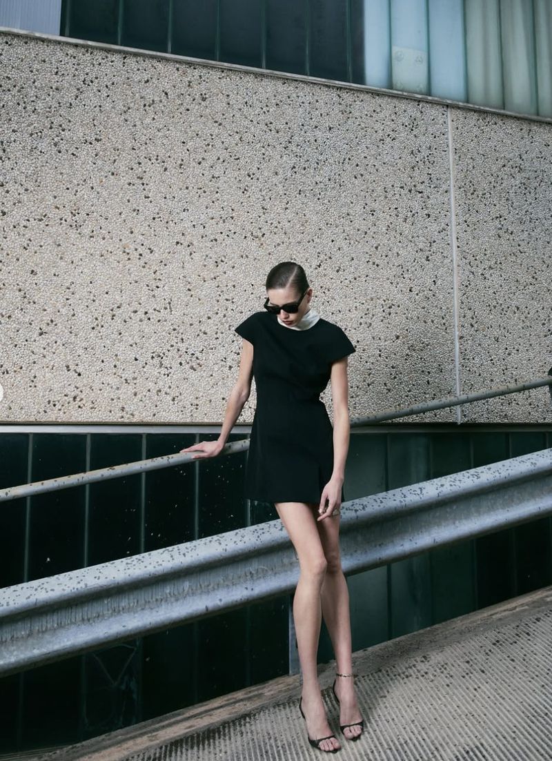 Woman in black dress and sunglasses poses on a ramp in an urban setting, leaning on a metal railing, creating a fashion-forward look.