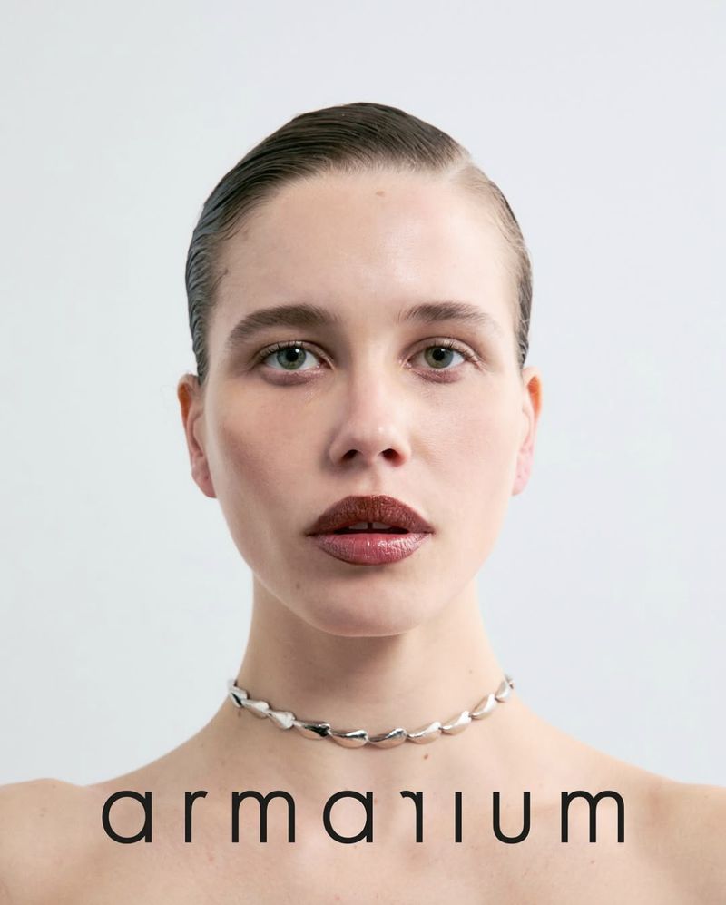 Close-up portrait of a woman with dark lipstick wearing a silver choker necklace against a neutral background, promoting fashion.