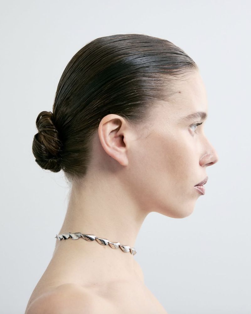 Profile of a woman with dark hair styled in a bun, wearing a silver necklace, posing in front of a white backdrop in a beauty shot.