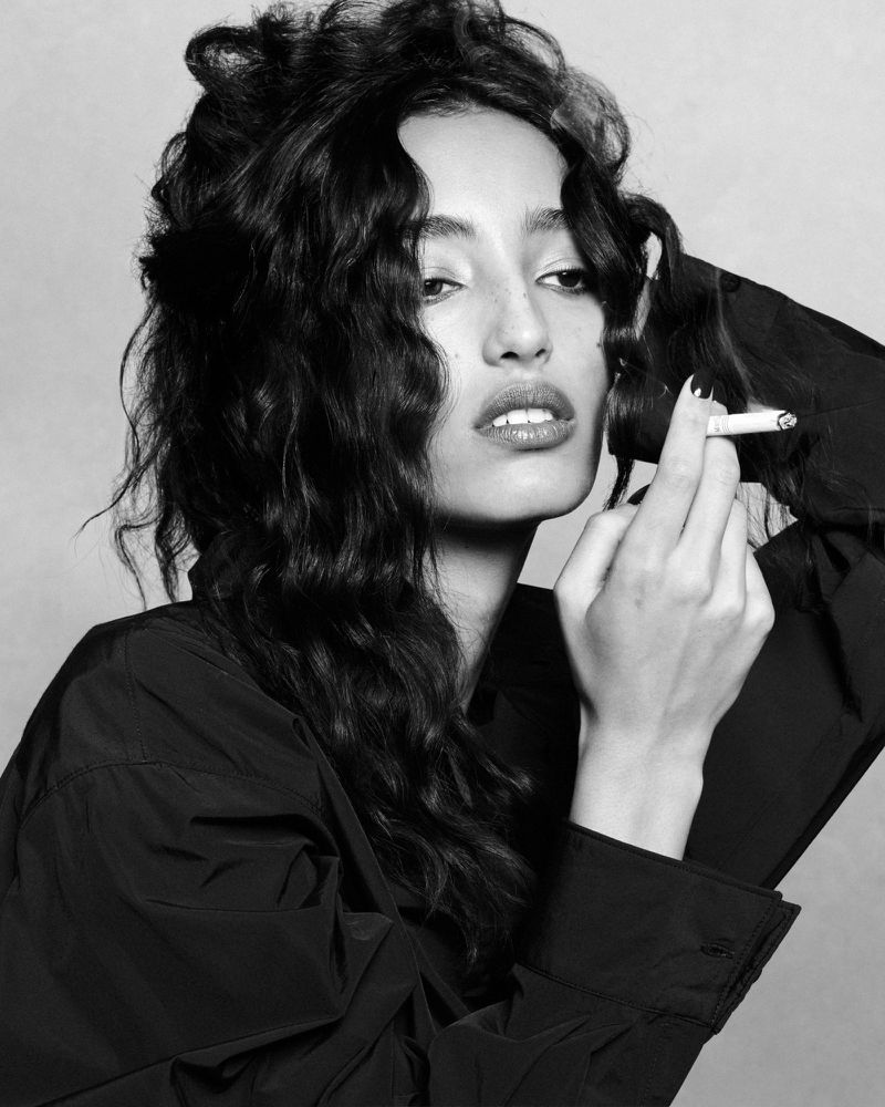Black and white portrait of model Mona Tougaard holding a lit cigarette wearing a dark shirt against a seamless background.