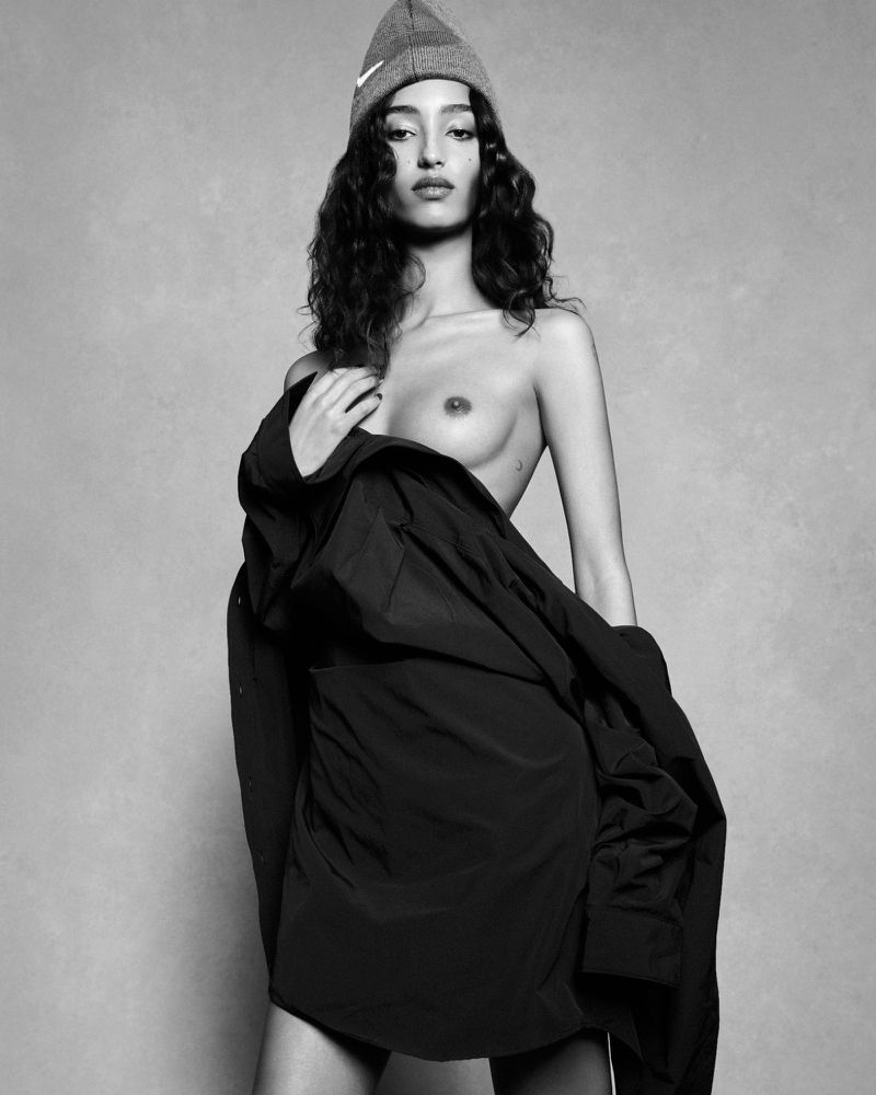 Model Mona Tougaard wearing a beanie and dark draped coat against a grey studio backdrop by Luigi & Iango