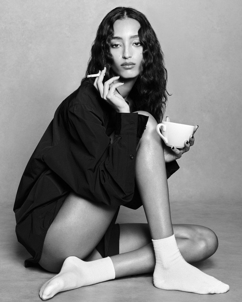 Model Mona Tougaard wearing a dark shirt and white socks holding a cup and cigarette against a plain studio background.