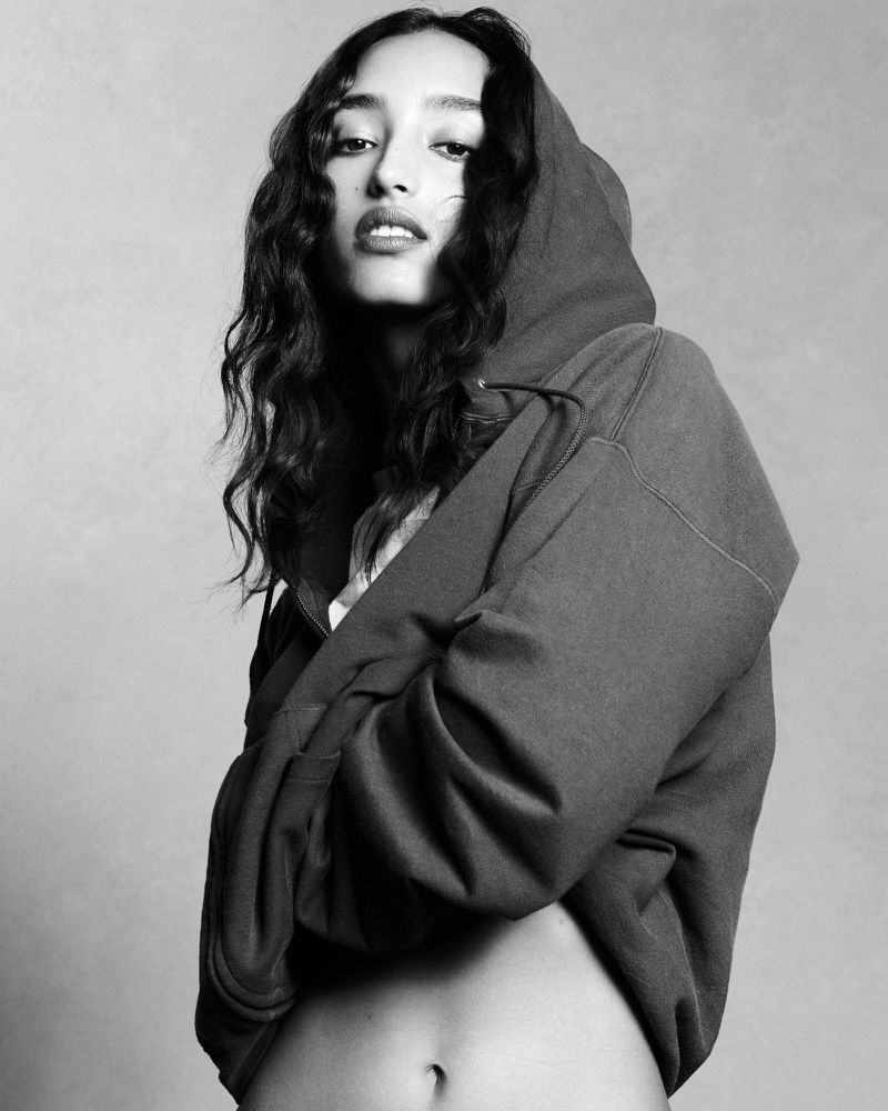 Model Mona Tougaard wearing a dark hooded sweatshirt against a textured grey studio background.
