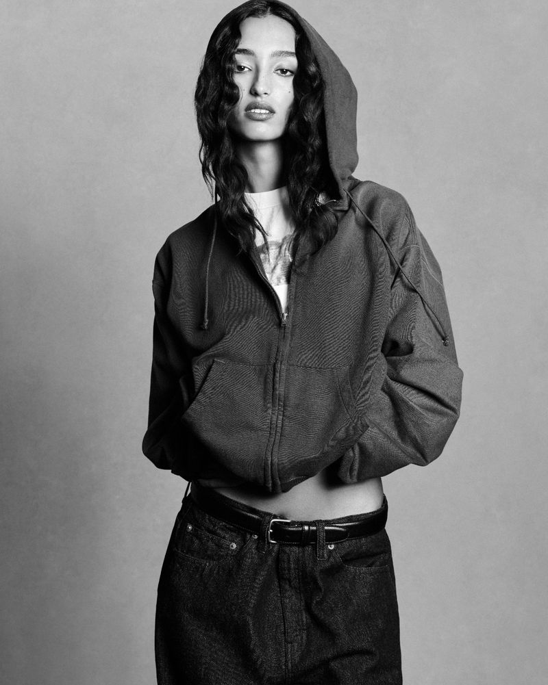 A woman in a hoodie poses in a studio portrait, standing in front of a gray background. She is wearing dark jeans and a belt.