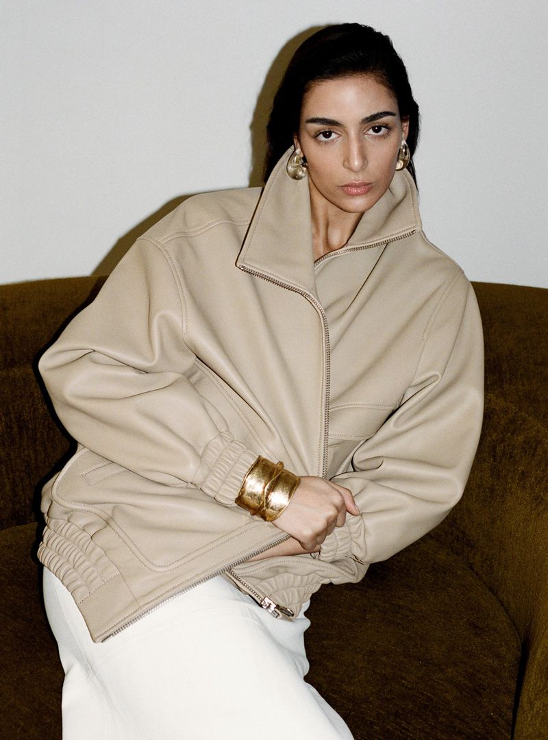 Model wearing a beige leather jacket and gold cuffs seated on a dark sofa against a plain white wall