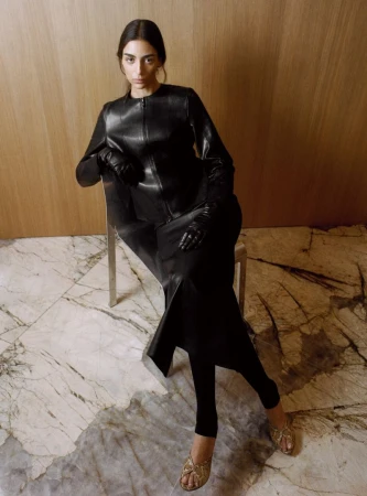 Model Nora Attal seated on a chair wearing a black leather jacket and skirt with gloves against a marble and wood paneling background