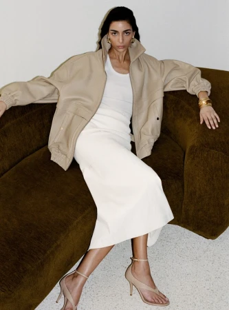 Model Nora Attal seated on an olive green sofa wearing a beige jacket and white skirt for Nour Hammour Spring Summer 26.