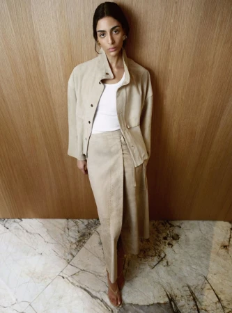 Model Nora Attal in a beige jacket and wrap skirt posing against a vertical wood panel wall.