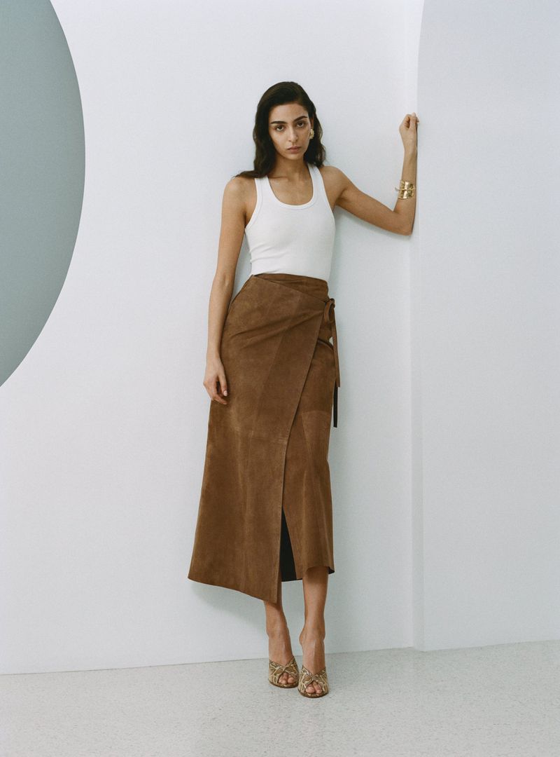 A woman poses in a white tank top and brown suede wrap skirt in a minimalist indoor studio, showcasing fashion and style.