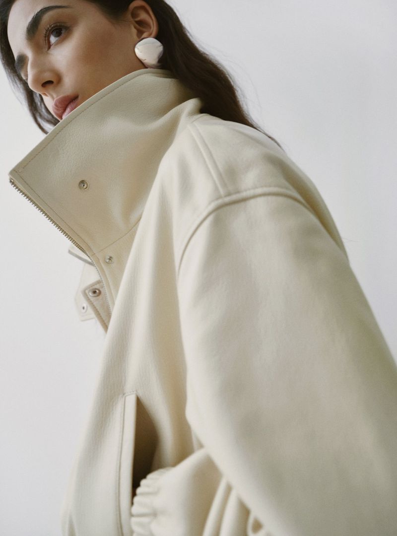 Model Nora Attal wearing a cream leather jacket with a high collar and white sculptural earrings against a plain background.