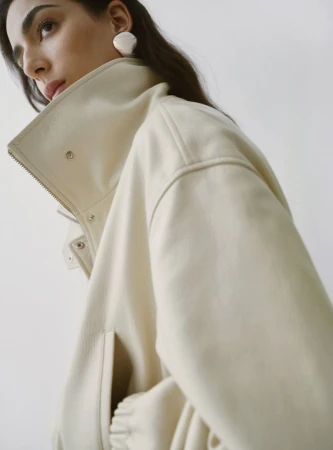 Model Nora Attal wearing a cream leather jacket with a high collar and white sculptural earrings against a plain background.