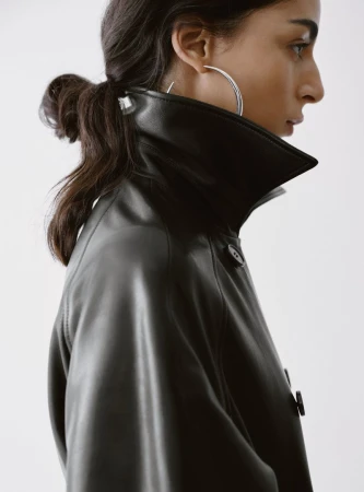 Profile view of Nora Attal wearing a black leather coat with a high collar and a large silver hoop earring against white seamless.