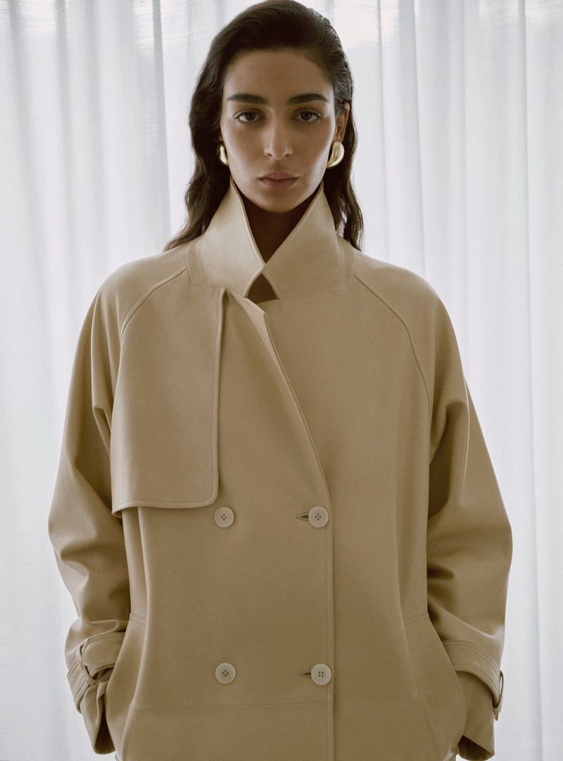 Model Nora Attal wearing a cream coat and gold earrings in front of white curtains for Nour Hammour Spring Summer 26.