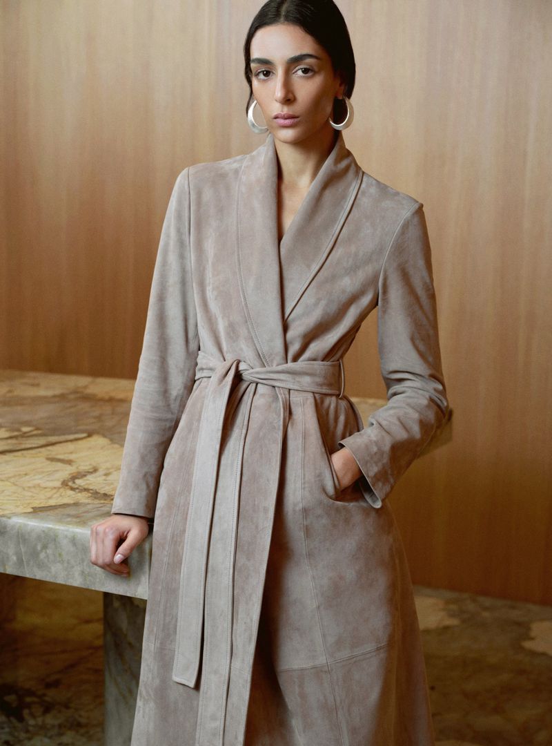 Model Nora Attal wearing a beige belted suede coat and silver hoop earrings against a warm wood paneled interior.