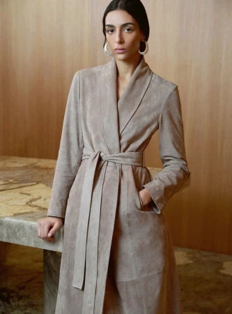 Model Nora Attal wearing a beige belted suede coat and silver hoop earrings against a warm wood paneled interior.