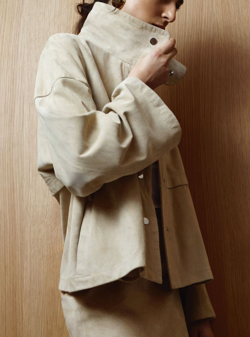 Model wearing a neutral-toned high-collar jacket against a vertical wood grain background.