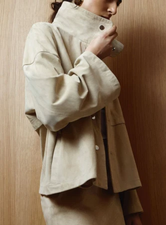 Model wearing a neutral-toned high-collar jacket against a vertical wood grain background.