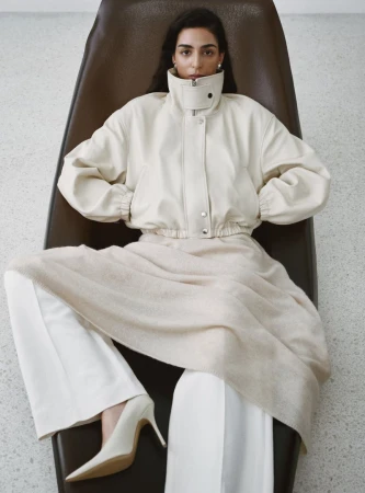 Model Nora Attal in a cream leather jacket and white trousers reclines on a dark leather lounge chair.