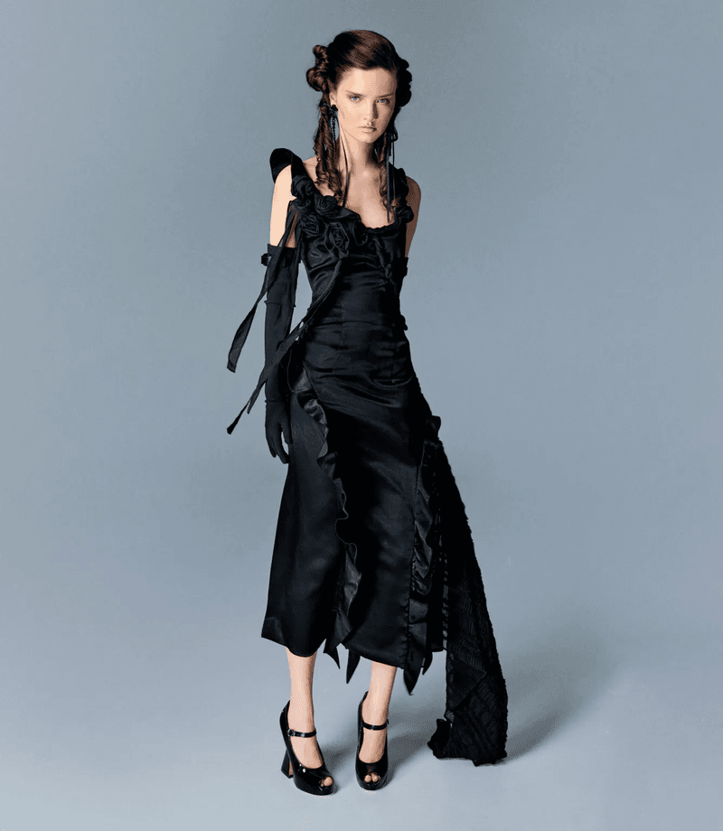 Model wearing a black ruffled dress and gloves posing against a plain grey background for an editorial project.
