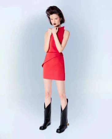 Model in a red minidress and tall black boots holding her arms across her chest against a pale gradient background.