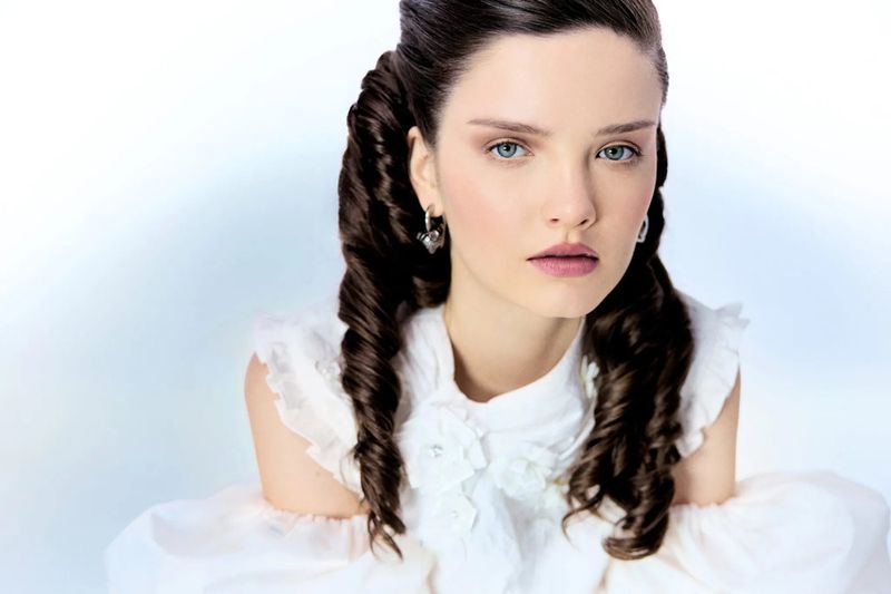 Portrait of a woman with elaborate dark coiled hair wearing a white ruffled top against a clean white background