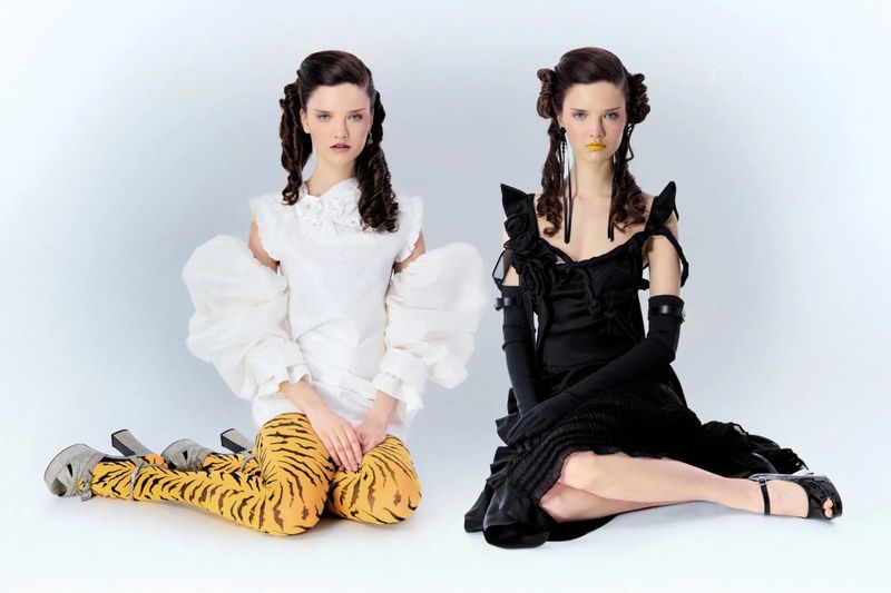 Two female models with elaborate braided hairstyles posing in contrasting white and black outfits against a white backdrop.