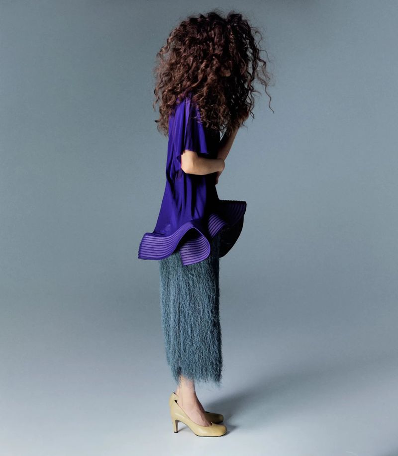 Model with voluminous curly hair wearing a purple top and textured grey skirt against a neutral grey background
