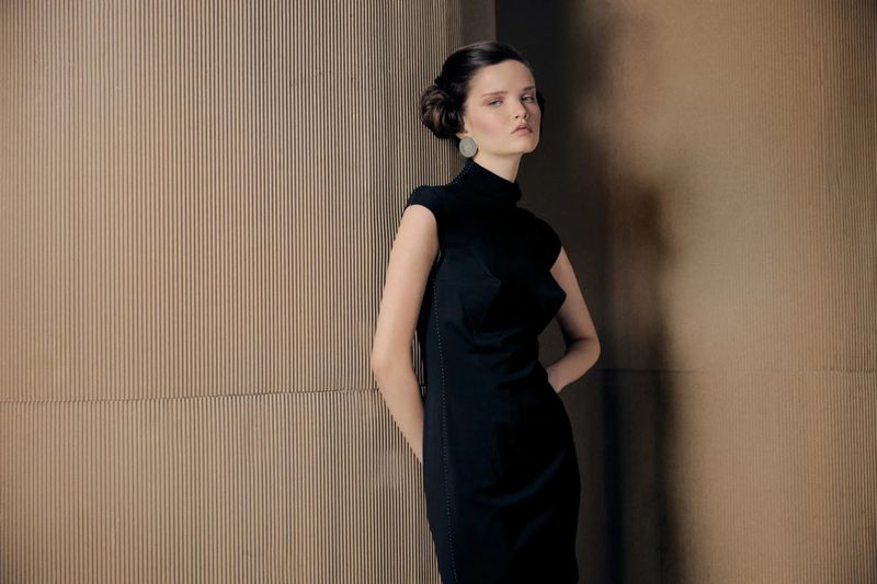 Model in a black sleeveless dress standing against a corrugated textured wall in an editorial fashion portrait.