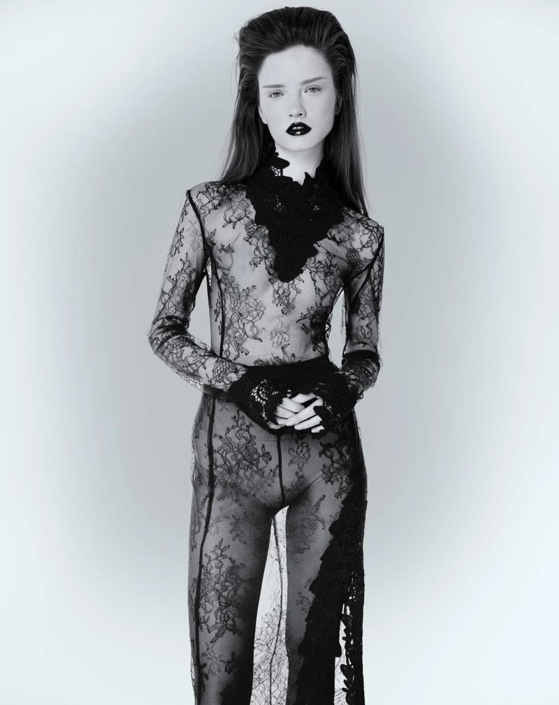 Model in sheer black lace bodysuit and dark lipstick standing against a plain light background for Elle Lithuania editorial