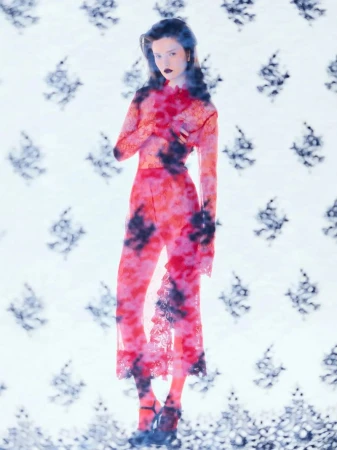 Model in a sheer red lace patterned jumpsuit standing against a light background with dark floral motifs for Elle Lithuania.