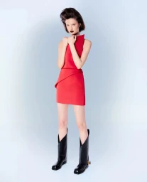 Model in a red minidress and tall black boots holding her arms across her chest against a pale gradient background.