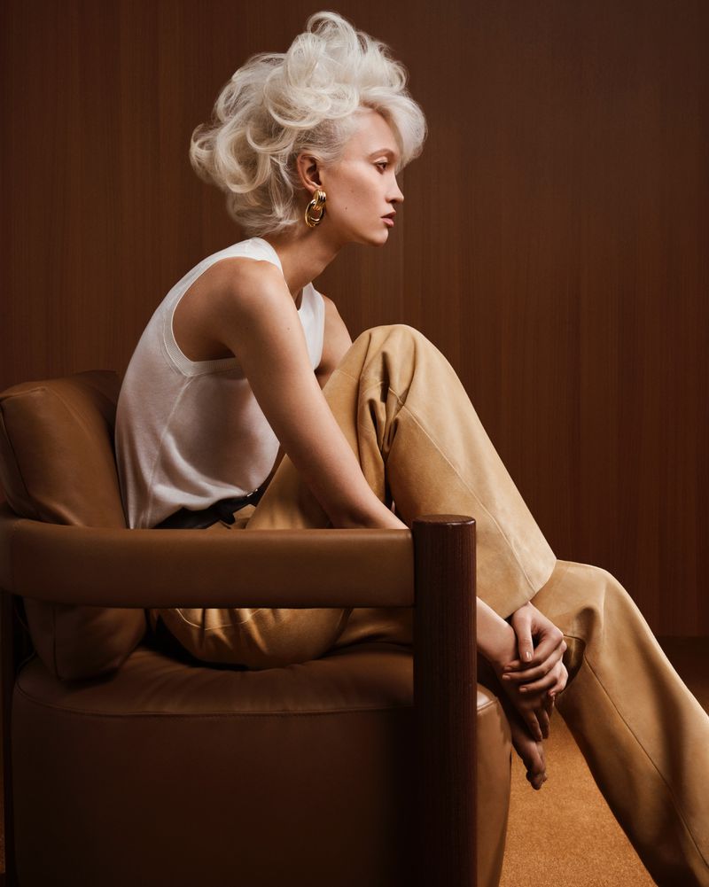 Model Patricia Zajac seated in a leather armchair wearing a white tank top and camel trousers for Grazia Italy.
